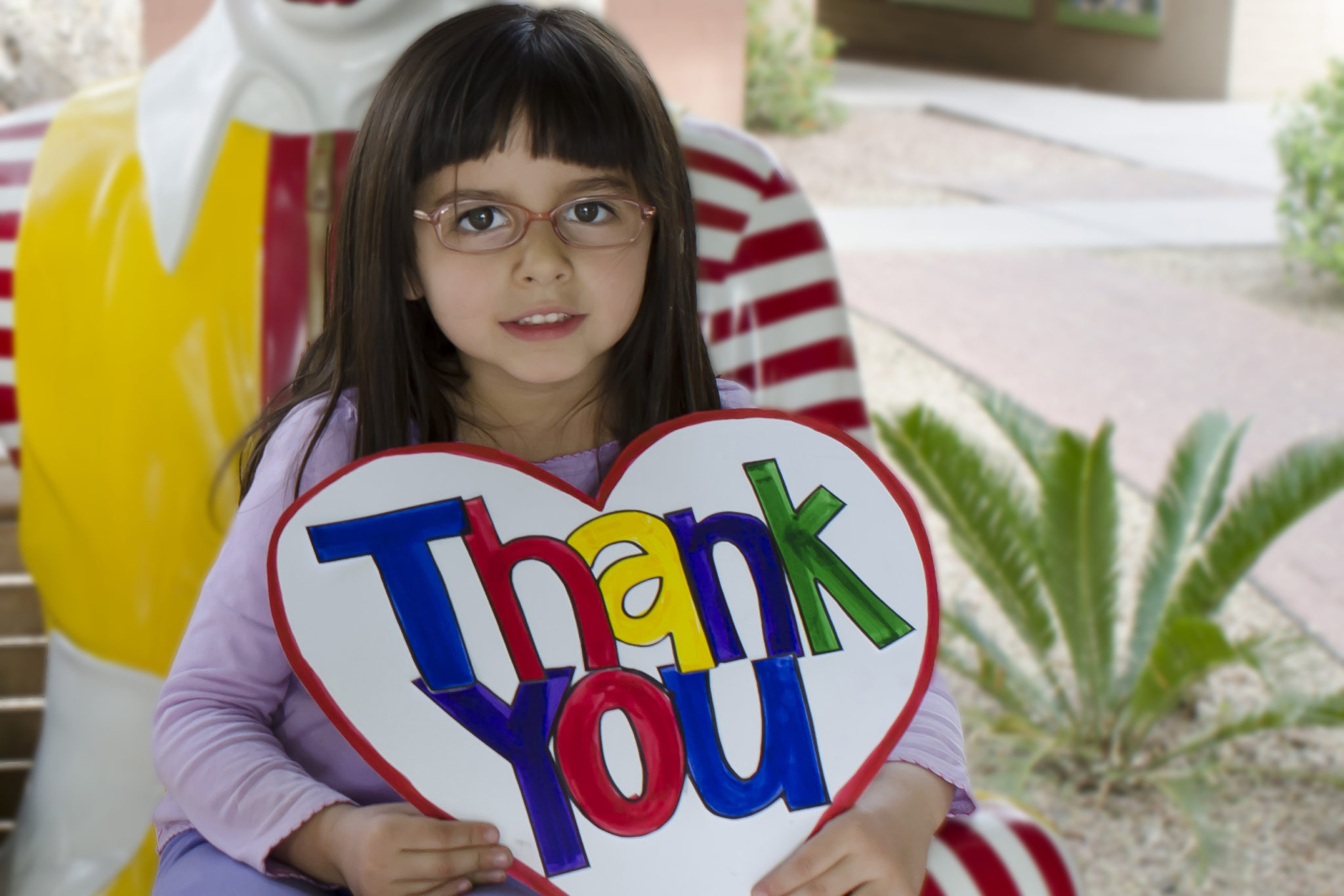 Little girl sitting on the Ronald McDonald bench holding a sign that says "thank you"